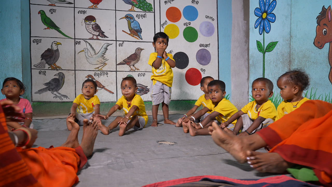 Children seated on a mat beneath a bird chart at an anganwadi centre, one child standing