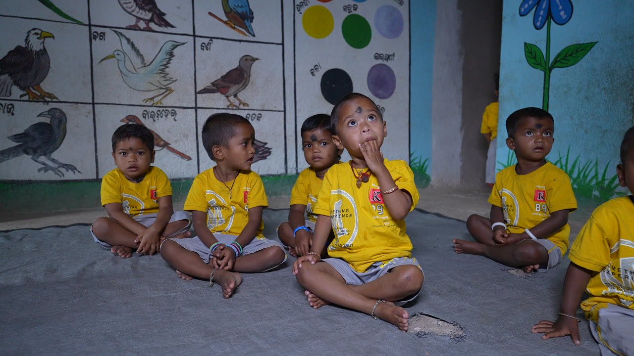 Children in yellow t-shirts sitting cross-legged along the anganwadi wall