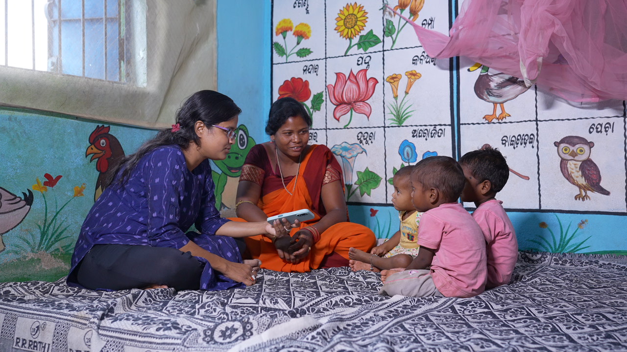 A field worker showing a phone screen to a mother and three children on a mat, beside a hand-painted flower and bird mural