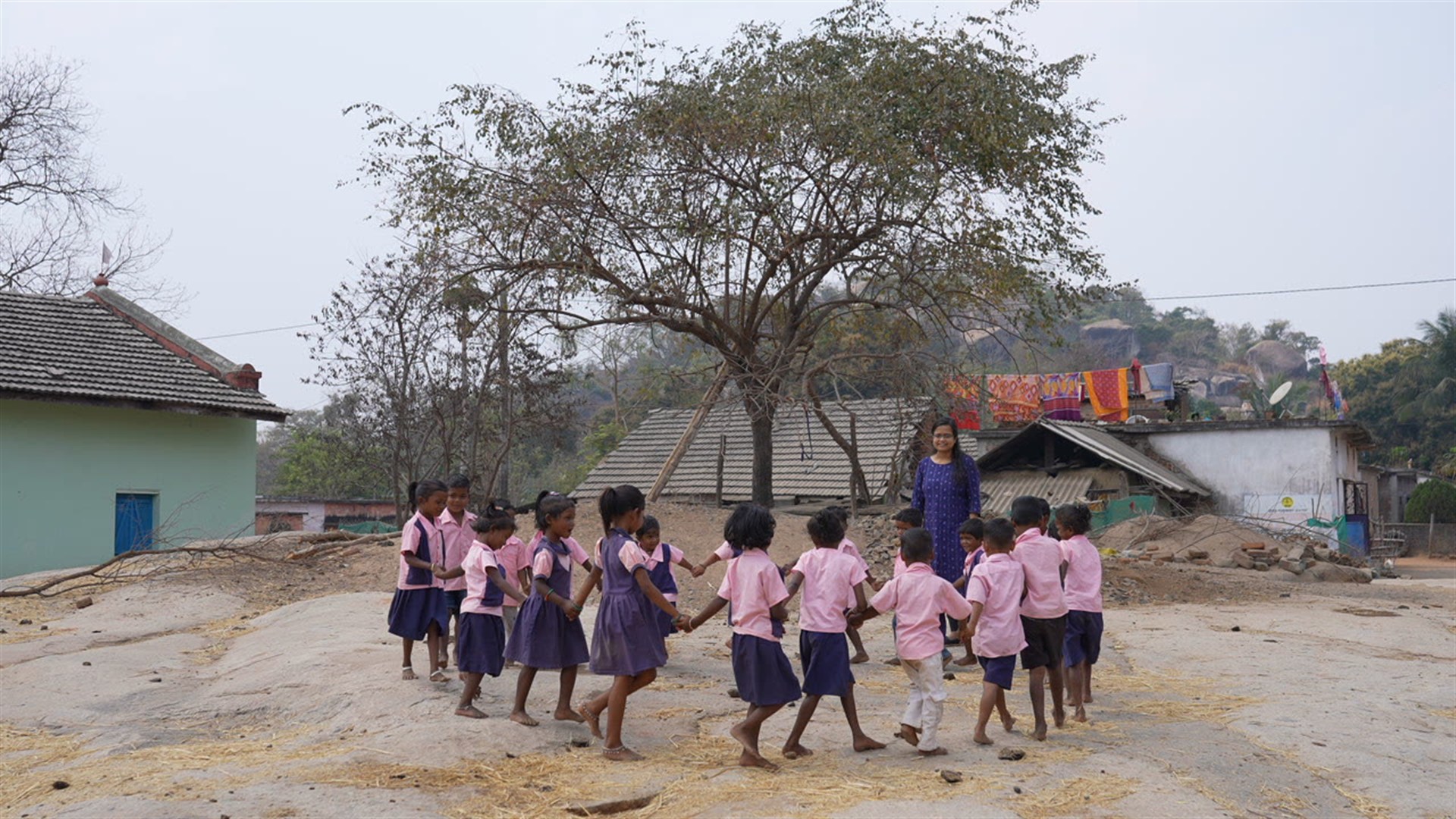 Village preschoolers in pink and purple uniforms holding hands in a circle game on open rock, their teacher standing among them, with tiled-roof houses and hills behind